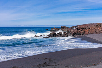 Fototapeta na wymiar The bright contrast of the white sea foam of the waves rolling onto the shore with the sand and black solidified lava.