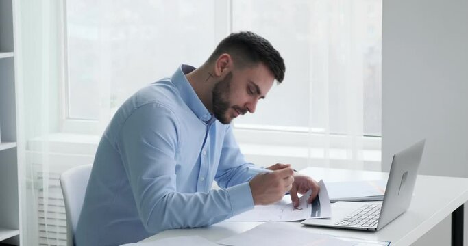Businessman with a beard sitting in an office, working. He is carefully reviewing documents that are spread out on his desk, which feature detailed graphs and statistics. He making notes with a pen.