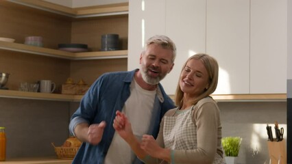 Middle-aged wife dance with senior husband adult couple listen music having fun dancing funny fooling around together in cozy kitchen cooking fresh salad romantic relations carefree moving spouses