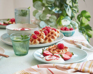 summer table with waffles served with yogurt, fresh raspberries and strawberries, refreshing beverages glasses and green leaf decoration