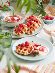Summer table with waffles, raspberries and yogurt, fresh drink and green leaves decoration.
