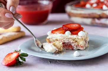 Italian traditional dessert tiramisu with strawberries. One piece of cake on a plate, woman hand holding fork, ingredients, cake over pink background
