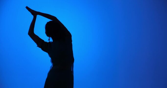 Silhouette of a talented young Indian dancer, dressed in a sari, as she moves gracefully against a blue background. Her skillful movements are sure to inspire awe and admiration.