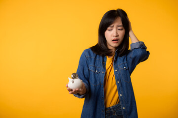 Fototapeta na wymiar Unhappy asian young woman wearing yellow t-shirt denim shirt pulling digital coin crypto currency out of piggy bank isolated on yellow background. Payment digital money debt financial concept.