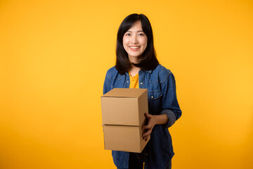 Portrait asian young woman wearing yellow t-shirt and denim shirt holding parcel box isolated on yellow studio background, Delivery courier and shipping service concept.