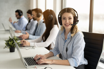 Friendly telephone operator woman with headset having conversation with client, showing excellent communication skills and attentive customer service, surrounded by group of colleagues at call center. - obrazy, fototapety, plakaty
