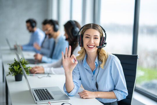 Friendly call center agent woman gesturing approval, showing the Ok sign to the camera while having a conversation with a satisfied client surrounded by supportive colleagues at office.