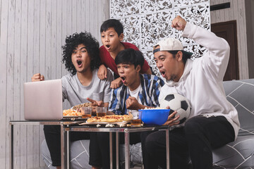 Happy Asian friends or football fans watching soccer on laptop and celebrating victory at home....