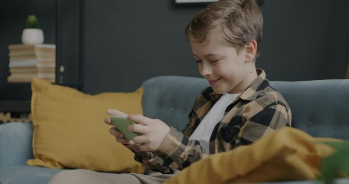 Excited boy having fun with smartphone playing video game winning laughing sitting on couch at home. Gaming and modern gadgets concept.