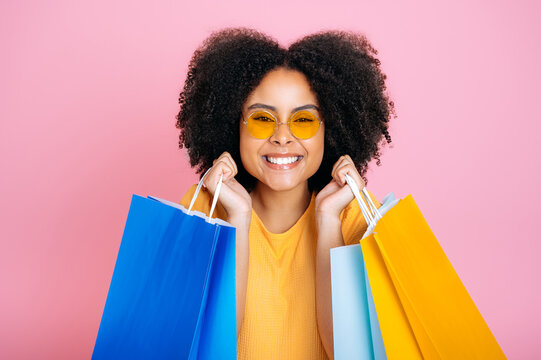 Close-up of a positive gorgeous excited curly haired brazilian or african american trendy girl, holding shopping paper bags, looking and smiling at camera, standing over isolated pink background