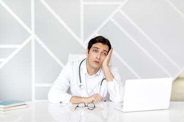 Close up portrait of tired male doctor sitting at the desktop and working on laptop in the office...