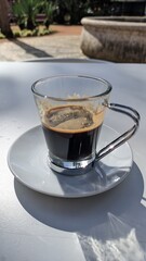 Small glass cup of black coffee with a metal handle in a white plate on the terrace
