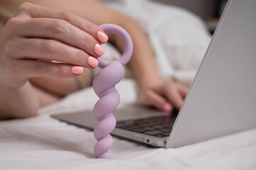Naklejka premium Woman holding lilac anal beads next to laptop while lying on bed. 