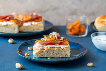Focaccia with prosciutto, soft cheese stracciatella, dry apricots and pistachios on a blue plate, ingredients, others sandwiches backside over dark blue and grey background