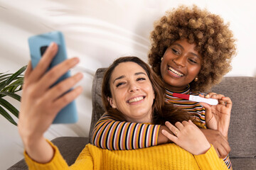 Diverse female couple taking a selfie with their positive pregnancy test in a sofa. Happy lesbian...