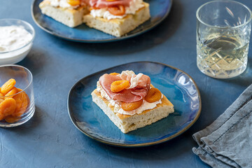 Focaccia with prosciutto, soft cheese stracciatella and dry apricots on a blue plate, grey napkin, drink, ingredients and others sandwiches backside over dark blue background