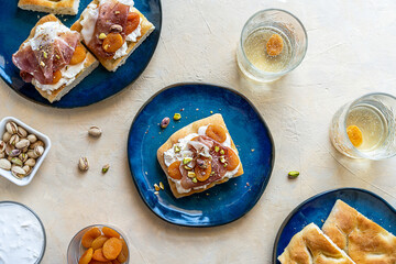 Focaccia sandwiches with prosciutto, soft cheese stracciatella, dry apricots and pistachios on blue plates, ingredients, fresh drink in glasses over light sand background
