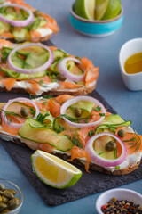 Open sandwiches with salted salmon, cream cheese, onions, capers, cucumber and microgreens on slate  board with white kitchen table background. Seafood. Healthy food. Close up. Copy space
