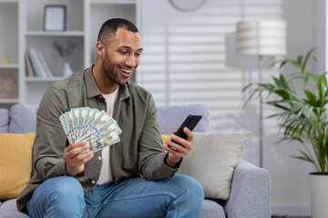 African american young man sitting on sofa at home and holding fan of cash money and dollars, using...