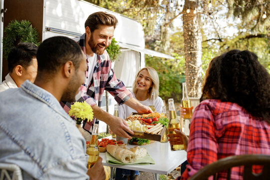 A cheerful multinational company is having fun on a picnic. Multisex friends sitting at the table outdoors celebrating eating and drinking drinks.