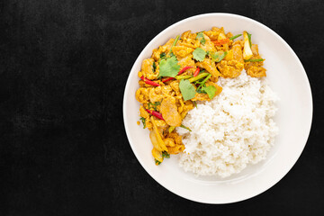 Stir Fried Chicken with Curry Powder beside Steamed Rice in simply white ceramic plate on dark tone texture background, top view, Gai Pad Pong Karee, Thai food