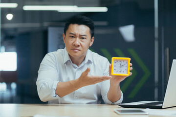 Portrait of a young Asian male businessman sitting in the office at the desk and showing the clock...