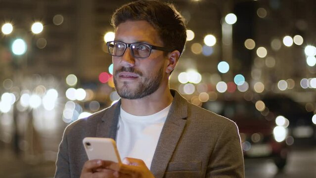 Businessman in City Center Street Uses Phone Texting Thinking Scrolling Tapping. Young Handsome Breaded Man Using Smart Phone. Technology And Success Concept. Traffic Night Slow Motion.