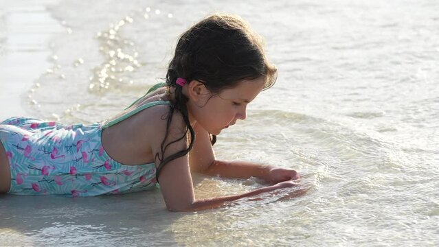 Little girl in blue bikini laying on white sand on beach of Caribbean Sea playing with waves on sunset light. Holidays and inspiration. Slow motion. High quality 4k footage
