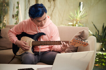 Young man following tutorial on laptop when learning playing guitar at home