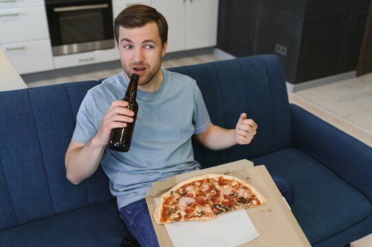 Eats pizza while watching TV show. Man with beard indoors.