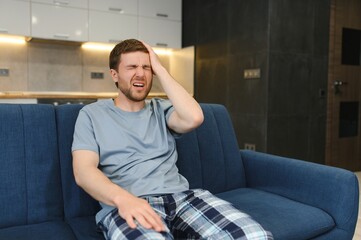 Unpleasant pain. Sad unhappy handsome man sitting on the sofa and holding his forehead while having headache.