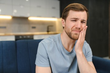 Toothache. Unhappy upset caucasian sad man sits on the couch at home, holds his hand near his...