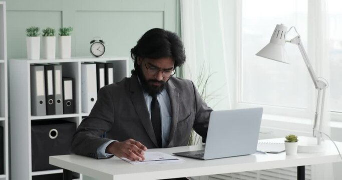 An Indian businessman wearing glasses and suit with tie sits in his office, reviewing documents on his laptop and cross-referencing information to ensure accuracy and efficiency.