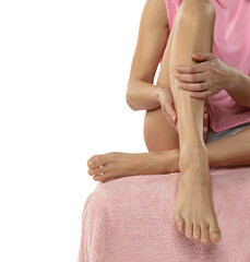Young woman sitting on bed and applying cream onto her leg after depilation on a white background