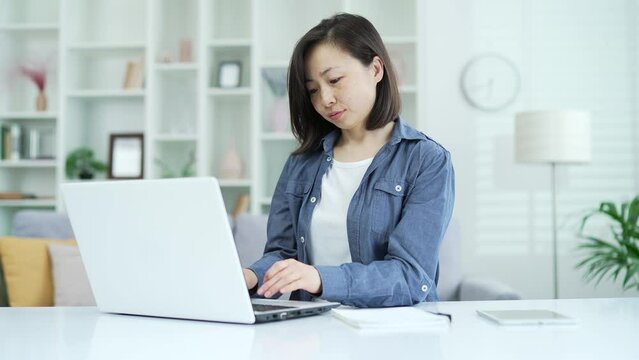 Adult asian businesswoman typing on laptop while sitting in home office. Female freelancer works on a computer on a project, chats online with a client, writes an email message or does online shopping