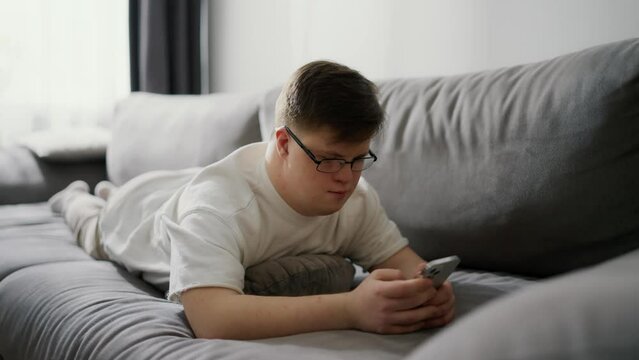 Smiling man with down syndrome using cellphone while resting on couch at home