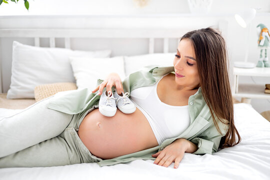 A pregnant woman with a big open belly holds socks and dreams of having a baby. The expectant mother is waiting and preparing for the birth of a child in a bright bedroom
