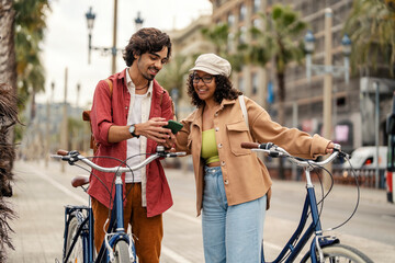 A cheerful tourist are standing on city street with their bikes and using online app. - obrazy, fototapety, plakaty