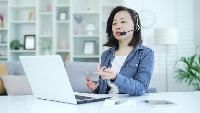 Confident asian woman in wireless headset talking on video call using laptop while sitting in home office. A female psychologist conducts an online consultation, communicates remotely with a patient