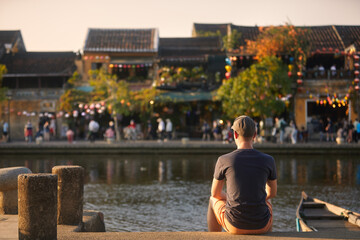 Rear view of man siting on waterfront in old town. Street of ancient city decorated with traditional lanterns, Hoi An, Vietnam.