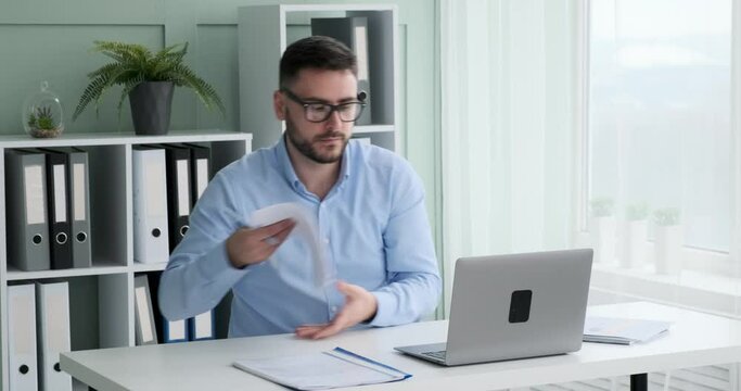 Businessman with a beard and glasses is hard at work in his office. He's sitting at his desk, laptop open and pen in hand, carefully reviewing important documents that showcase graphs and statistics.