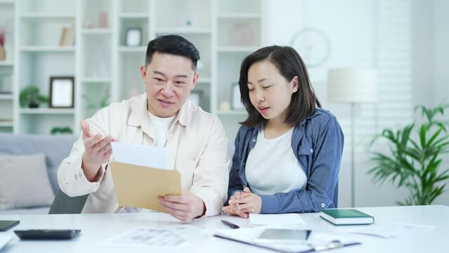 Happy asian couple celebrating success reading a letter with great news while sitting in home office. Excited smiling joyful husband and wife satisfied with pleasant notification, showing yes gesture