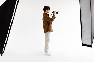Full length shot of young photographer man holding professional photocamera, standing on white...