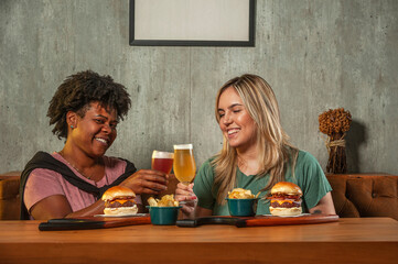 Brazilian girls holding a glass of beer with burger sitting at restaurant fast food table - Girls having lunch break at cafe bar - Life style concept with girls holding beer.