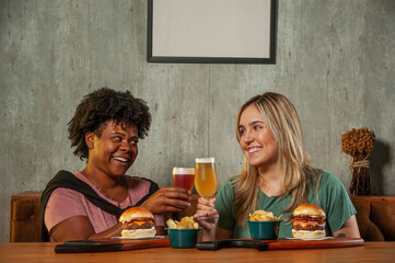 Brazilian girls holding a glass of beer with burger sitting at restaurant fast food table - Girls having lunch break at cafe bar - Life style concept with girls holding beer.
