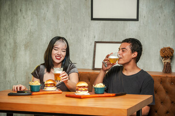 Happy couple drinking beer sitting at pub restaurant fast food table with burger and potato chips.