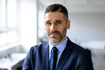 Fototapeta premium Closeup portrait of bearded middle aged businessman posing at office, man manager in suit and tie looking at camera