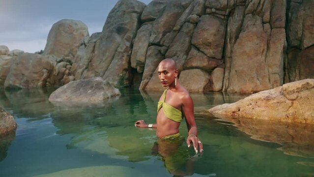 LGBTQIA black person in luxury dress walks, turns back at camera in water of natural ocean pool. Gender fluid ethnic stylish model wearing jewellery dressed in open outfit in tropical rocky lake.