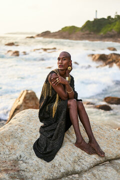 Epatage lgbtq black man poses on scenic ocean beach looks at camera demonstrates jewellery . Androgynous ethnic fashion model in posh dress looks at camera, sits on stone in