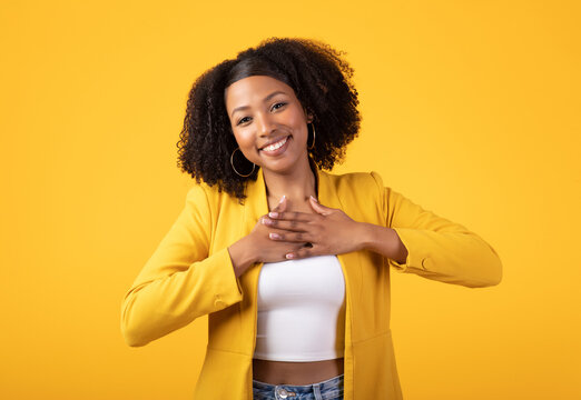 Thank you. Thankful black lady pressing hands to chest and smiling, standing posing over yellow studio background